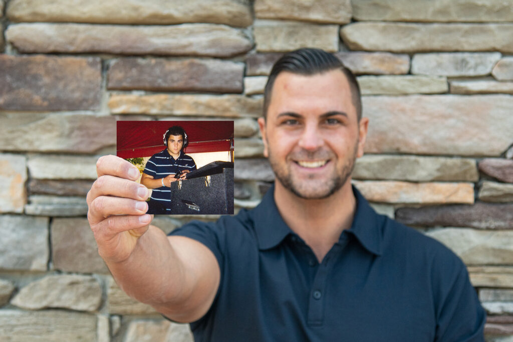 Image of  Jason McConnell smiling in a blue polo shirt, holding a phot of him deejaying while standing in front of a white stone wall