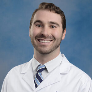James' headshot with blue background in white coat and blue striped tie