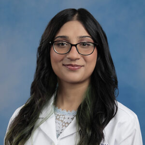 Fizza's headshot with blue background in white coat and long dark hair with glasses