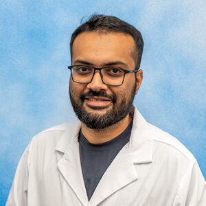 Dr Sheheryar's smiling headshot with blue background and white coat