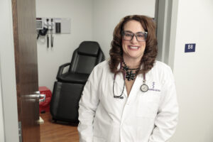 Dr. Linda Thomas-Hemak standing outside patient room door frame with white coat and glasses