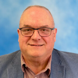 Mark's smiling headshot with blue background and glasses and grey blazer