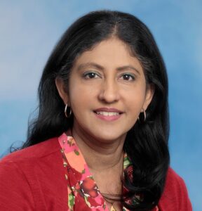 Dr. Manju Mary Thomas smiling headshot with blue background and red top and long dark hair