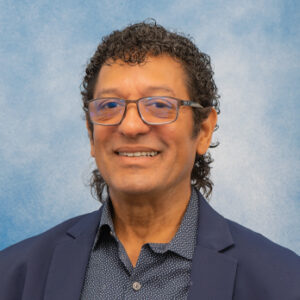 Maximo's smiling headshot with blue background and navy blazer with glasses