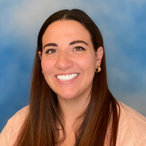 Maria's smiling headshot with blue background and peach top and long brown hair