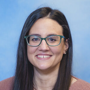 Shauna's smiling headshot with blue background and green glasses