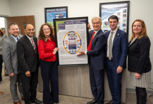 A group photo of Dr Oz, Rob Bresnahan, Dr Thomas, Dr Sheth, Brian Ebersole, and Jenn Walsh standing in a line holding a framed poster of our GME safety-net consortium