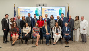 Group photo of Dr Oz with Dr Thomas and other Wright Center employees, and board members in two rows with some seated and some standing and a screen in behind them with our wright center ten year vision