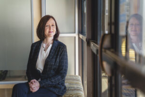 Nicole Flynn posing by window on a bench in checked dark blazer
