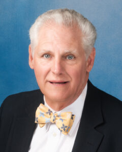 John P. Kearney's headshot on blue background with bowtie and black suit coat