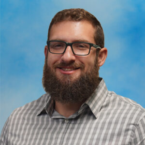 Portrait of Corey Kucker wearing glasses, smiling in front of a blue background.